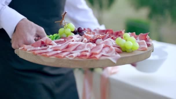 Primo piano colpo di cameriere in uniforme mettendo grande bordo di legno con fette di prosciutto sul tavolo da banchetto che si prepara per la festa di nozze di lusso, servo organizzare tavolo per gli ospiti di nozze all'aperto. Ristorazione — Video Stock