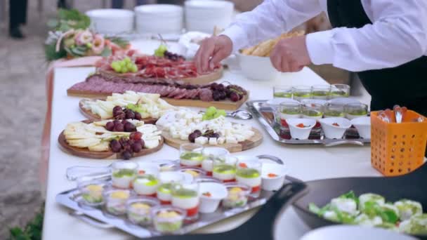 Restauracja kelnerzy przygotowuje stolik w formie bufetu na przyjęcie weselne, służący umieszczenie sztućce dla zaproszonych gości na stole. Srebrne tacki i drewniane deski pełne mięsnych przekąsek, przystawek i kanapek — Wideo stockowe