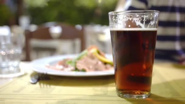 Sklenice osvěžujícího červeného piva na rozmazaném stolním pozadí restaurace, středomořská pochoutka na bílém talíři tradičního místního baru, hospody nebo restaurace. Filé z uzeného lososa s raketovým salátem a — Stock video
