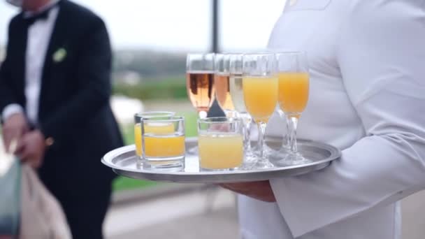 Camarero en uniforme blanco sosteniendo bandeja de plata con copas de champán, zumbido de vino blanco y jugo fresco preparado para los huéspedes. Bebidas de lujo en fiesta de celebración, servicio de catering y banquete — Vídeos de Stock