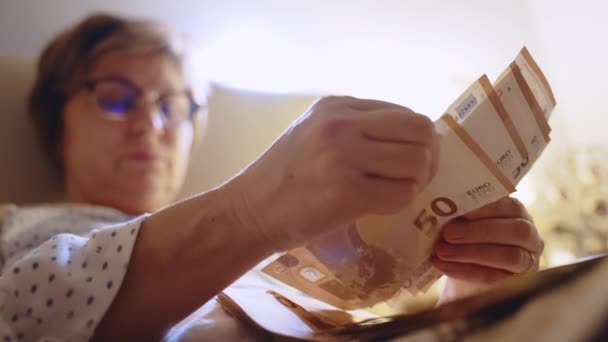 Сконцентрированная женщина среднего возраста в очках, лежащих в постели и подсчитывающая бумажную валюту евро от двух пачек, грустная женщина, подсчитывающая свой ежемесячный доход, откладывающая деньги на оплату счетов и имущественный кредит — стоковое видео