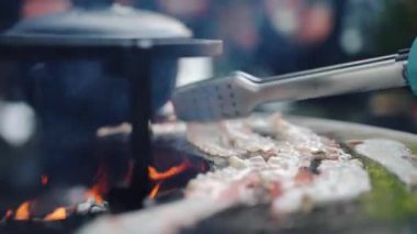 Şefin eldiven ve eldiveni yakın plan çekimi. Yemek maşası kullanarak BBC sigara içme ızgarasında ızgara domuz pastırması dilimleri, hafta sonu arkadaşlarıyla yapılan bbq partisi. Tuzlu dilimlenmiş domuz pastırması.