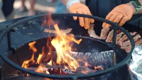 Fumante de churrasco tradicional para assar variedade de carne para festa de piquenique bbq, queimando fogo no meio de fumante redondo e grelhando fatias de bacon crocante ao redor das chamas de fogo. Chef em luvas — Vídeo de Stock