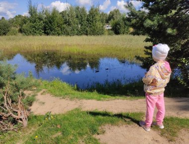 Küçük bir kız ördeklerin yüzdüğü göle bakıyor. Güneşli havalarda çekilmiş bir fotoğraf, bir insanın sırtından. Belarus 'un manzara ve çizimleri.