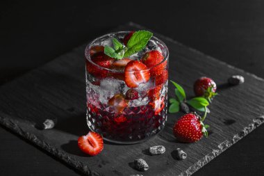Soğuk, ev yapımı, cam bardakta buzlu çilekli limonata, koyu arkaplanda nane yaprakları ve meyveler..