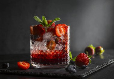 Soğuk, ev yapımı, cam bardakta buzlu çilekli limonata, koyu arkaplanda nane yaprakları ve meyveler..