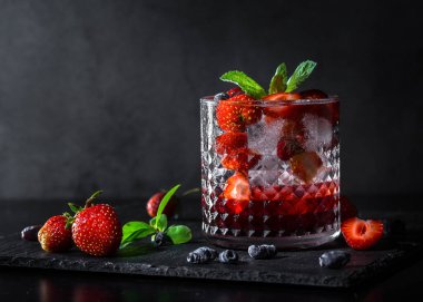 Soğuk, ev yapımı, cam bardakta buzlu çilekli limonata, koyu arkaplanda nane yaprakları ve meyveler..