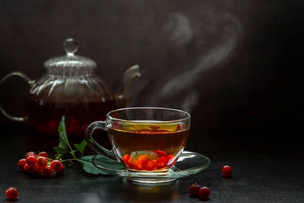 Hot tea with hawthorn in a transparent cup and a teapot with tea on a dark background.
