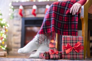 Kış çoraplarıyla kaplı kadın bacakları ekoseli oturma ve şöminenin yanındaki koltukta rahatlama ve aile için Noel ağacı paketleme kutuları. Alt görünüm.