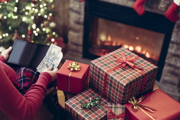 圣诞节 身穿毛衣 头戴平板电脑 头戴美元搜索礼物的女人坐在壁炉边和圣诞树旁的桌子旁 — 图库照片