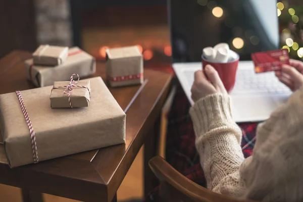 ラップトップを使用してクレジットカードを保持している女性は 暖炉やクリスマスツリーの近くのパッケージギフト ココアとマシュマロのカップでテーブルに座って注文をする オンラインショッピングのコンセプト — ストック写真