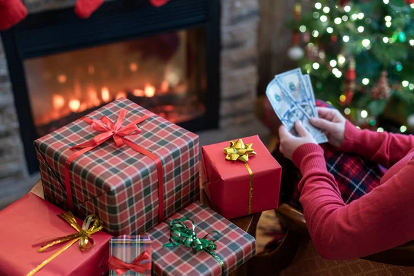 一个女人坐在圣诞树旁 壁炉旁和礼品盒旁 数着美国的美元 在圣诞节的时候花钱 — 图库照片