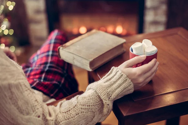 クリスマスツリーと暖炉の近くの木製テーブルの上に古いレトロな本とホットココアとマシュマロのカップを持つ女性の手 トーン — ストック写真