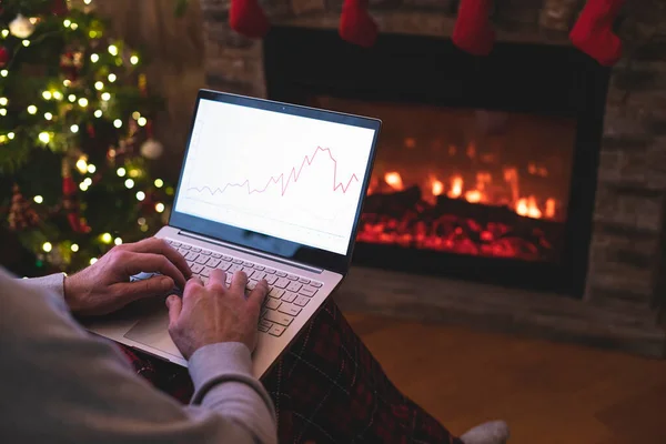 Man freelancer in santa claus hat working on laptop with graphs and charts on screen sitting near christmas tree and fireplace.