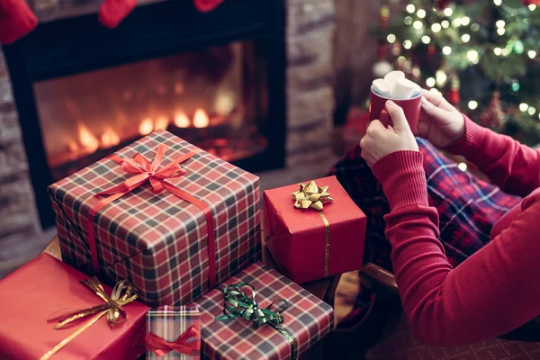 ホットチョコレートと溶かしたマシュマロの赤いマグカップを持つ女性クリスマスツリーと暖炉の近くに座って手作りのクリスマスプレゼントを梱包 — ストック写真