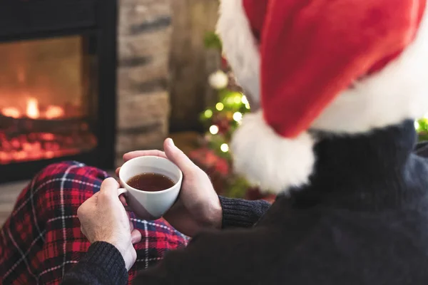 一位离婚男子坐在壁炉边 手里拿着一杯咖啡 戴着圣塔克洛斯帽喝茶 冬天的夜晚还在炉边取暖 — 图库照片