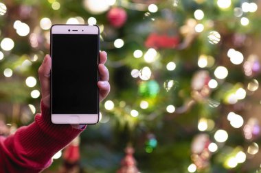 Cep telefonu tutan ve arka planda Noel ağacı ışıkları olan bir kadın. Tatillerde çevrimiçi hizmet kavramı.