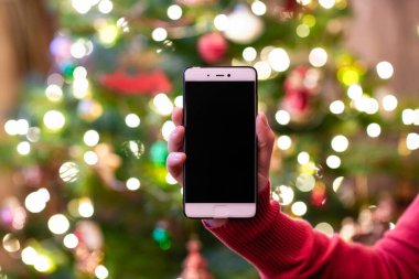 Cep telefonu tutan ve arka planda Noel ağacı ışıkları olan bir kadın. Tatillerde çevrimiçi hizmet kavramı.