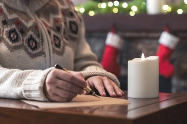 Bir kadın Noel şöminesinde dolma kalemle dilek listesi yazıyor. Ampul süslemesi ve masa üzerinde mum var..