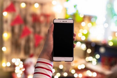 Cep telefonu tutan ve arka planda Noel ışıkları olan ekranı gösteren bir kadın. Tatillerde çevrimiçi hizmet kavramı.