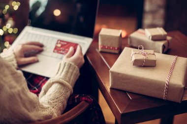 Noel zamanı. Elinde kredi kartı olan bir kadın sipariş vermek için dizüstü bilgisayar kullanıyor şömine ve yılbaşı ağacının yanında paketleme hediyesiyle oturuyor. Çevrimiçi alışveriş kavramı