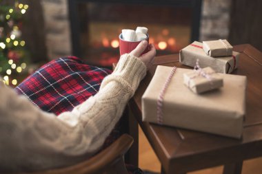 Kadın, aile için hediye paketlerini bitirdikten sonra ekose oturma ve rahatlama ve noel ağacını kakao ve marşmelovla kapladı.. 