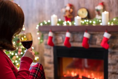 Arka planda cam kaplı ekoseli şarap içen bir kadın Noel arifesi ve köknar ağacının yanındaki koltukta oturuyor ve rahatlıyor..