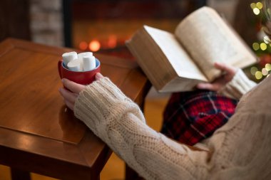 Woman hand with old retro book and cup of hot cocoa and marshmallow on wooden table near christmas tree and fireplace.  clipart