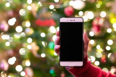 Cep telefonu tutan ve arka planda Noel ağacı ışıkları olan bir kadın. Selamlama için bir model. Kavram. 