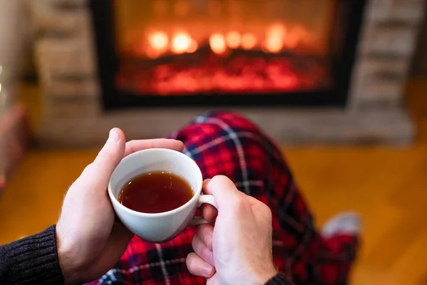 Man with cup coffee, tea in santa claus hat sitting and warming at winter evening near fireplace flame, covered christmas plaid