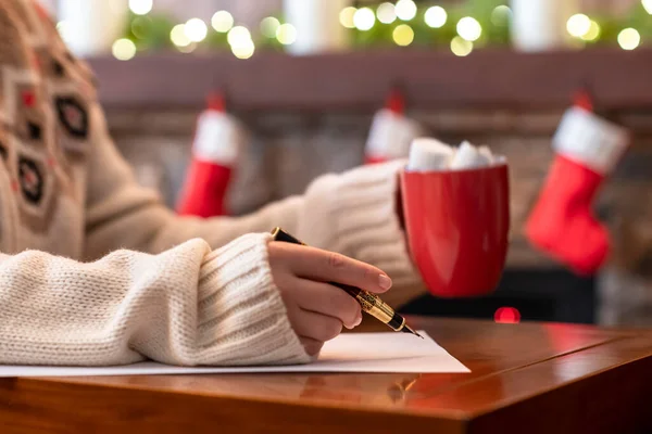 Woman writing wish list using fountain pen on sheet of paper at christmas fireplace with decoration of light bulbs drinking hot chocolate and marshmallow.