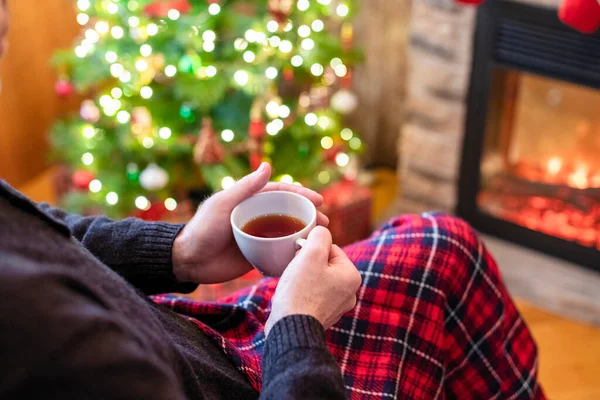 男人端着一杯咖啡 头戴圣爪帽喝茶 冬夜坐在壁炉边取暖 涂满了圣诞花纹 — 图库照片