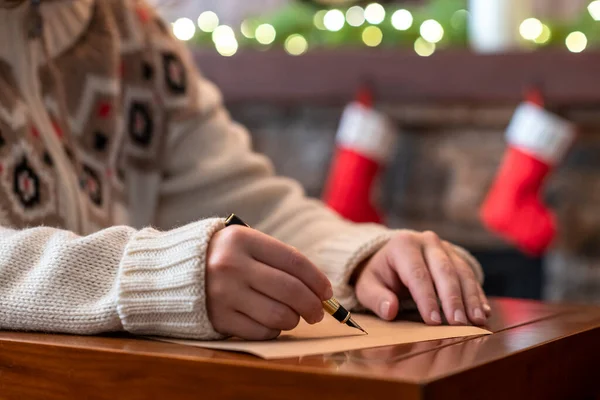 Hand of young woman with fountain pen and sheet of paper on background of christmas tree and fireplace with decoration of light bulbs. Concept of writing letter to Santa Claus