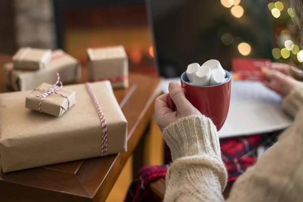 ラップトップを使用してクレジットカードを保持している女性は 暖炉やクリスマスツリーの近くのパッケージギフト ココアとマシュマロのカップでテーブルに座って注文をする オンラインショッピングのコンセプト — ストック写真