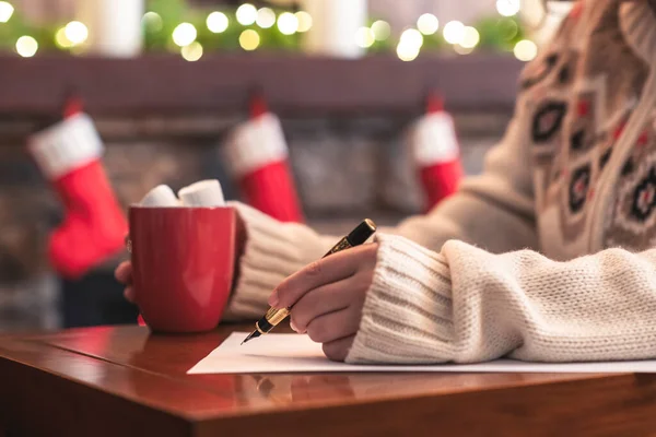 Woman writing wish list using fountain pen on sheet of paper at christmas fireplace with decoration of light bulbs drinking hot chocolate and marshmallow.