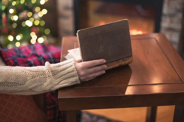 Woman hand with old retro book on wooden table near christmas tree and fireplace. Toned