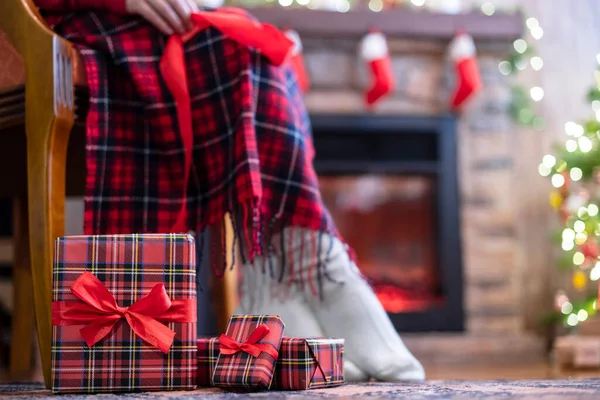 Woman legs in a winter socks covered plaid sitting and relaxation on armchair near fireplace and christmas tree pakking gift boxes for family. Bottom view.