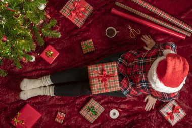 Dizlerinin üzerinde Noel hediyesi kutusu olan bir kadının en iyi görüntüsü. Noel ağacının yanındaki kabarık ekose üzerinde oturan aile için kadın paketleme hediyeleri. Düz yatıyordu.