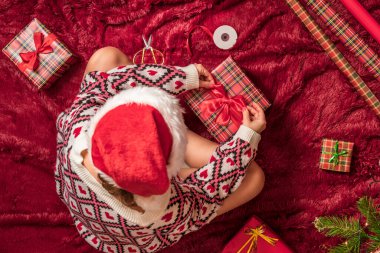 Kırmızı Noel Baba şapkalı bir kadının, Noel ağacının yanındaki kabarık ekose kumaşta oturan aile için Noel hediyesi kutularını paketlediği en iyi manzara. Düz yatıyordu.