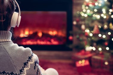 Kulaklıklı kadın kış akşamı şömine ateşi ve yılbaşı ağacının yanında oturup ısınıyor..