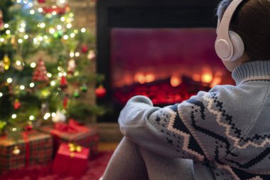 Yanlız üzgün kulaklıklı kadın kış akşamı şömine ateşi ve yılbaşı ağacının yanında oturup ısınıyor..