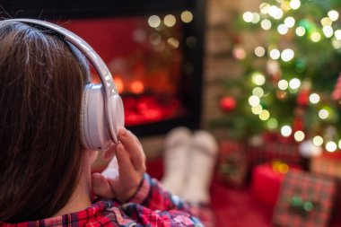 Kulaklıklı bir kadın kış akşamı şömine ateşi ve noel ağacının yanında oturup ısınıyor..