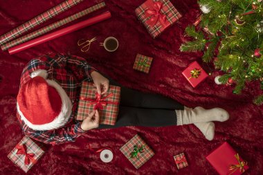 Dizlerinin üzerinde Noel hediyesi kutusu olan bir kadının en iyi görüntüsü. Noel ağacının yanındaki kabarık ekose üzerinde oturan aile için kadın paketleme hediyeleri. Düz yatıyordu.