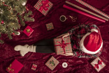 Dizlerinin üzerinde Noel hediyesi kutusu olan bir kadının en iyi görüntüsü. Noel ağacının yanındaki kabarık ekose üzerinde oturan aile için kadın paketleme hediyeleri. Düz yatıyordu.