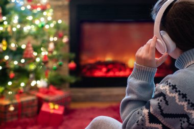 Kulaklıklı kadın kış akşamı şömine ateşi ve yılbaşı ağacının yanında oturup ısınıyor..