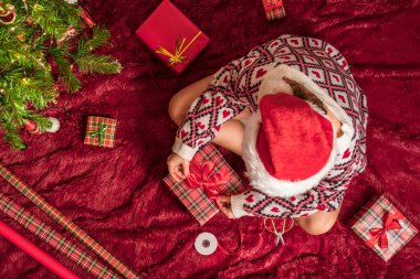 Kırmızı Noel Baba şapkalı bir kadının, Noel ağacının yanındaki kabarık ekose kumaşta oturan aile için Noel hediyesi kutularını paketlediği en iyi manzara. Düz yatıyordu.