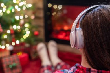 Kulaklıklı bir kadın kış akşamı şömine ateşi ve noel ağacının yanında oturup ısınıyor..