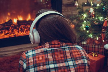 Kulaklıklı bir kadın kış akşamı şömine ateşi ve noel ağacının yanında oturup ısınıyor..