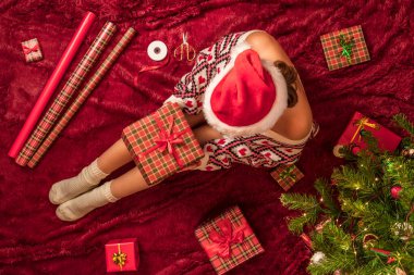 Dizlerinin üzerinde Noel hediyesi kutusu olan bir kadının en iyi görüntüsü. Noel ağacının yanındaki kabarık ekose üzerinde oturan aile için kadın paketleme hediyeleri. Düz yatıyordu.