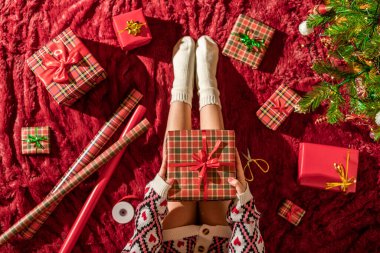 Dizlerinin üzerinde Noel hediyesi kutusu olan bir kadının en iyi görüntüsü. Noel ağacının yanındaki kabarık ekose üzerinde oturan aile için kadın paketleme hediyeleri. Düz yatıyordu.